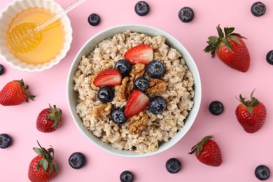 Photo of Tasty oatmeal with strawberries, blueberries and walnuts in bowl surrounded by fresh berries on pink background, flat lay