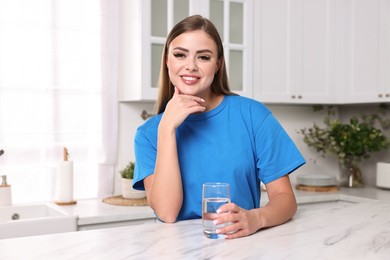 Happy woman with glass of fresh water at white marble table in kitchen