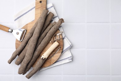 Photo of Raw salsify roots and peeler on white tiled table, top view. Space for text