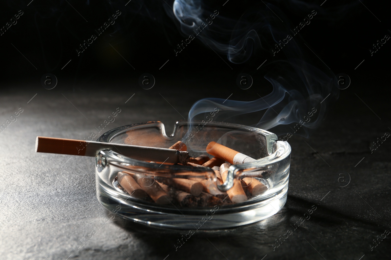 Photo of Smoldering cigarette in ashtray on grey table against black background