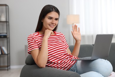 Photo of Happy young woman having video chat via laptop and waving hello on sofa in living room