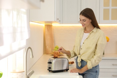 Young woman taking slice of bread from toaster in kitchen