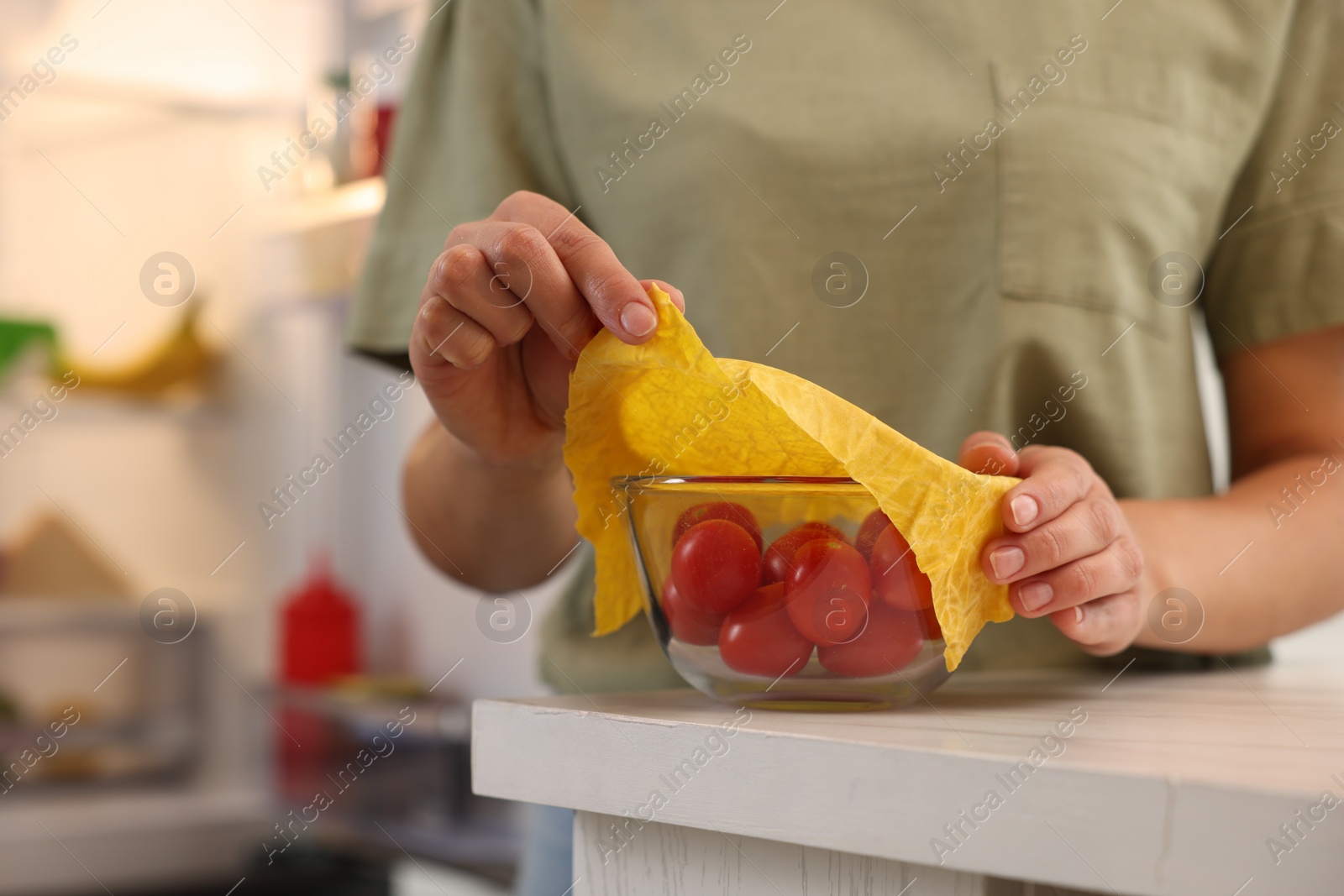 Photo of Woman covering bowl of fresh tomatoes with beeswax food wrap at table in kitchen, closeup. Space for text