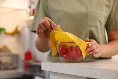 Woman covering bowl of fresh tomatoes with beeswax food wrap at table in kitchen, closeup. Space for text