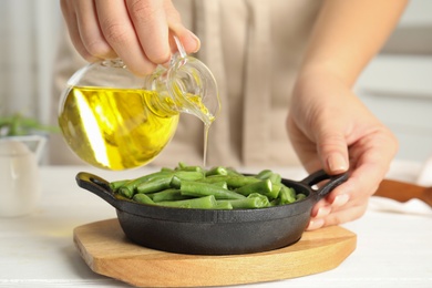 Photo of Woman pouring oil onto green beans at white wooden table, closeup