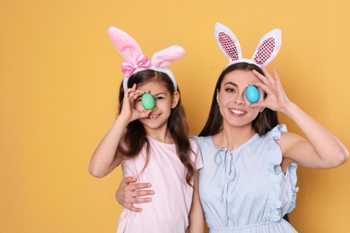 Mother and daughter in bunny ears headbands holding Easter eggs near eyes on color background