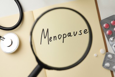 Notebook with word Menopause, pills and stethoscope on white table, view through magnifying glass