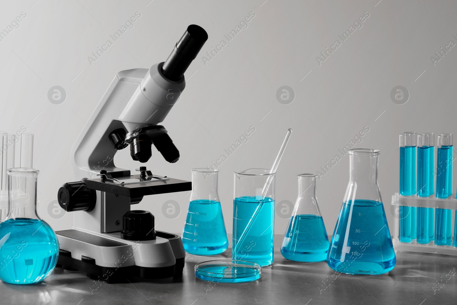 Photo of Different laboratory glassware with light blue liquid and microscope on table