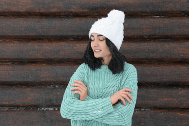 Young woman wearing warm sweater and hat near wooden wall. Winter season