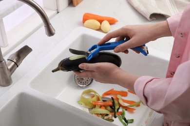 Photo of Woman peeling cucumber over kitchen sink with garbage disposal at home, closeup