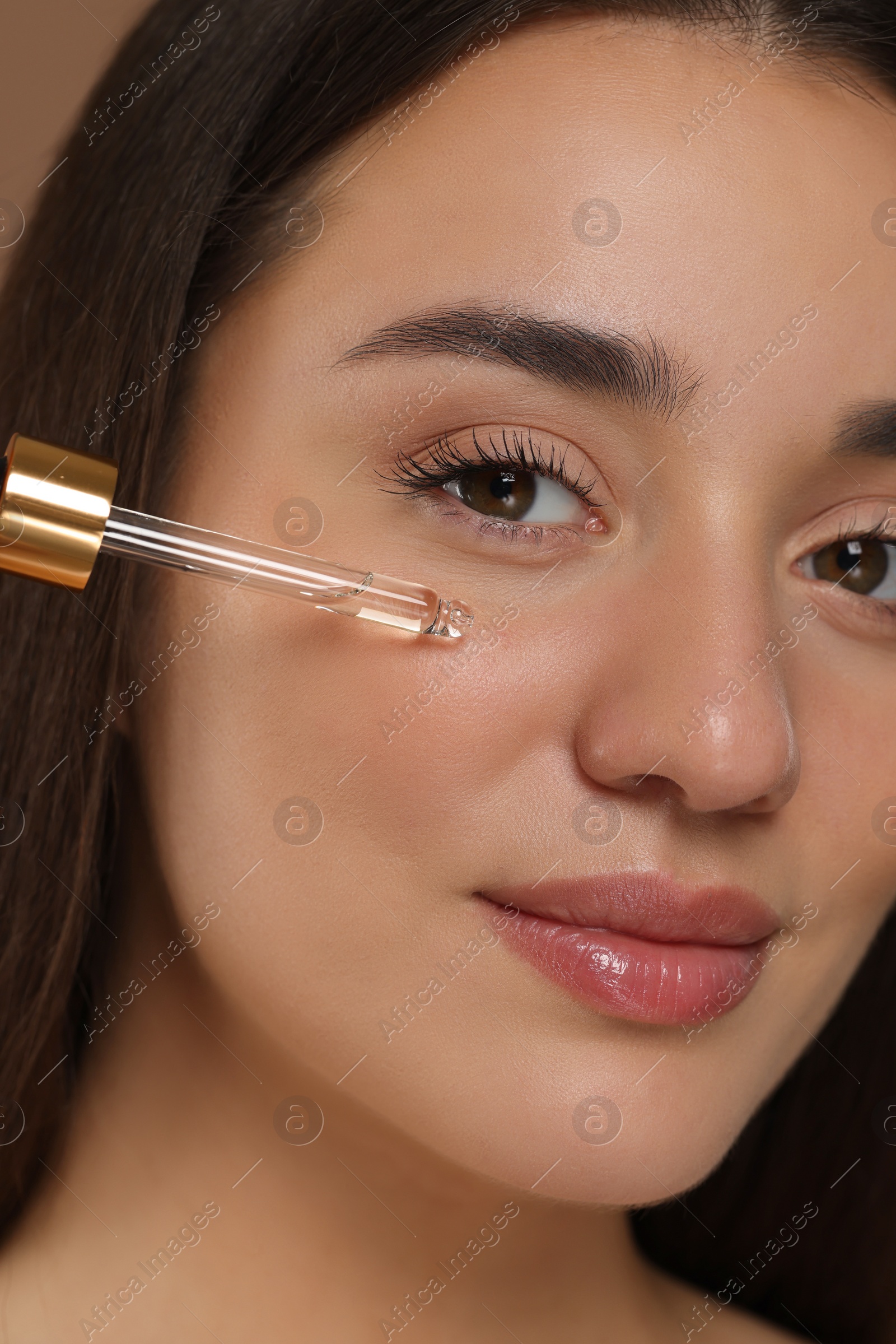 Photo of Beautiful woman applying essential oil onto face on brown background, closeup