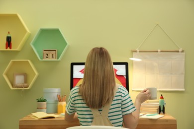 Photo of Woman sitting at wooden desk with computer near light green wall, back view. Interior design