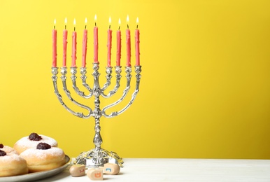 Photo of Silver menorah near sufganiyot and dreidels with symbols Nun, He, Pe, Gimel on yellow background, space for text