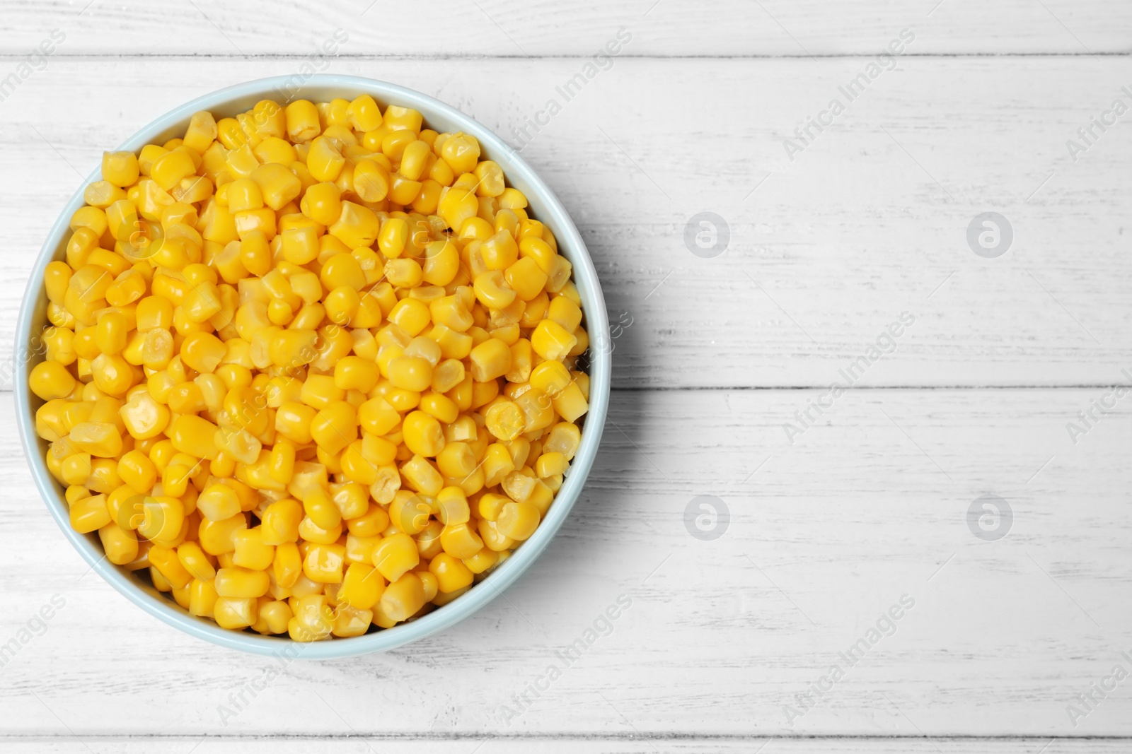 Photo of Delicious canned corn in bowl on white wooden table, top view. Space for text