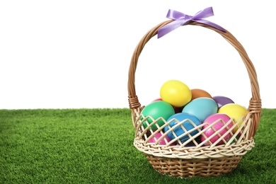 Photo of Wicker basket with bow and bright painted Easter eggs on green grass against white background. Space for text