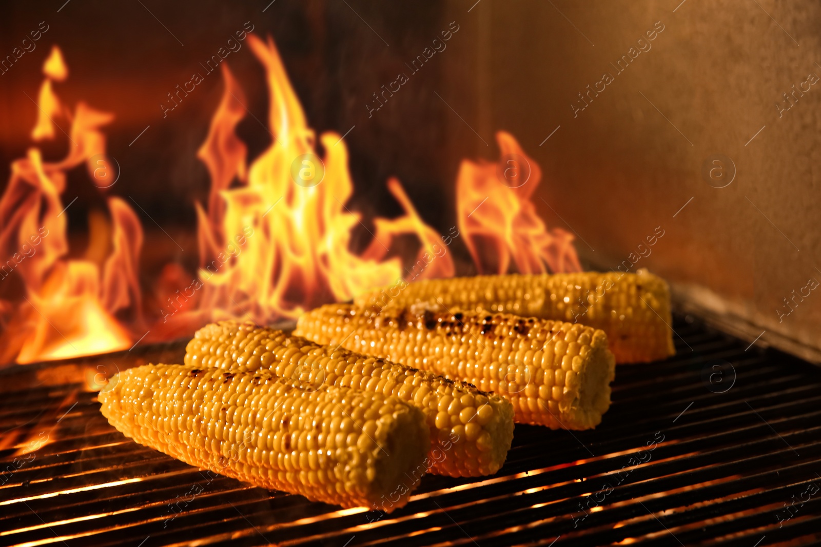 Photo of Cooking delicious fresh corn cobs on grilling grate in oven with burning firewood