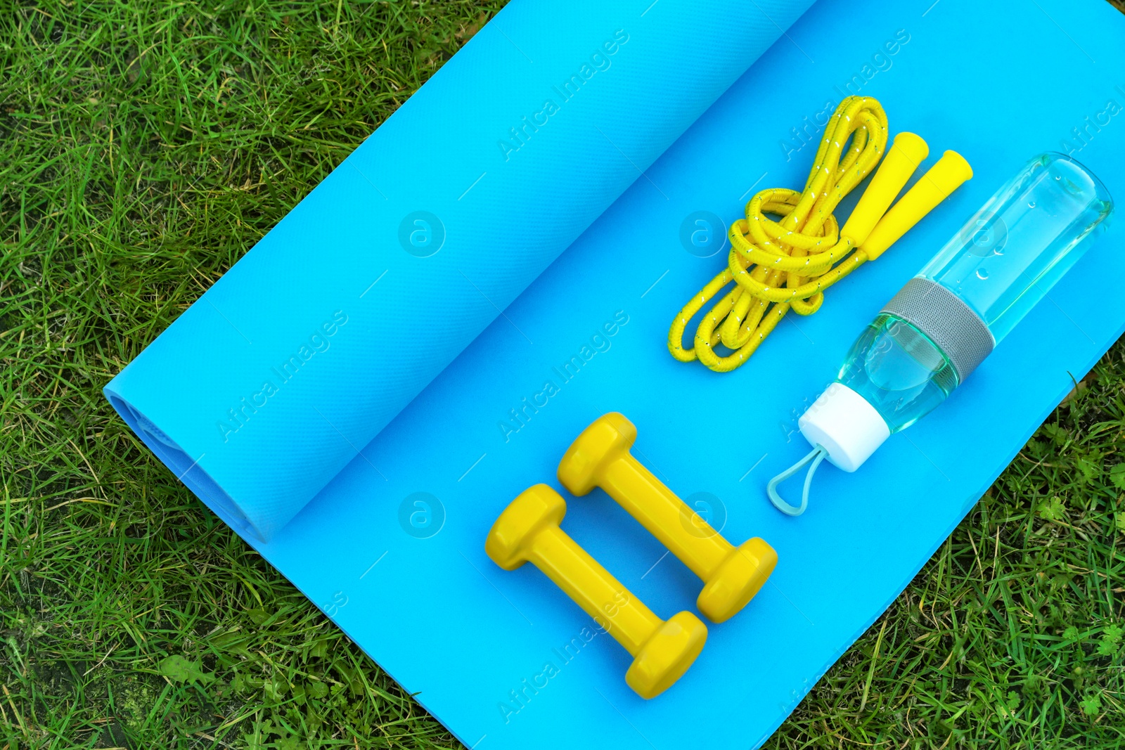 Photo of Skipping rope, dumbbells, bottle of water and fitness mat on green grass, flat lay. Morning exercise