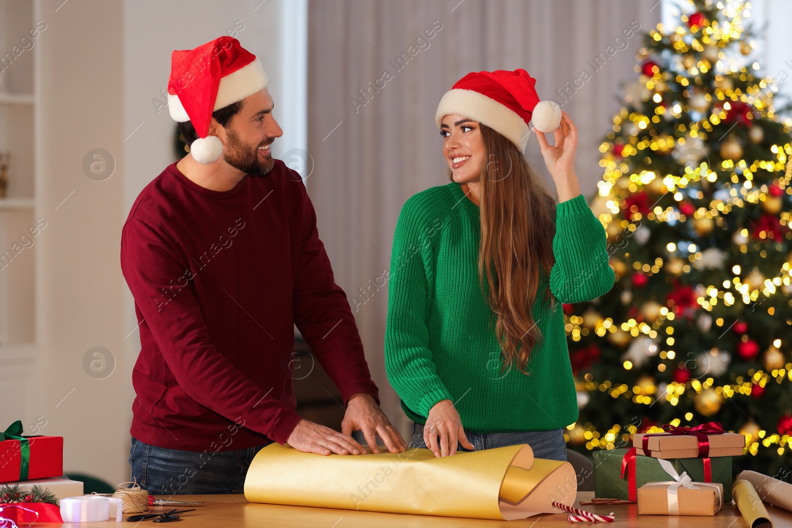 Photo of Happy couple in Santa hats decorating Christmas gift with wrapping paper at table in room