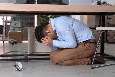 Scared man hiding under office desk during earthquake