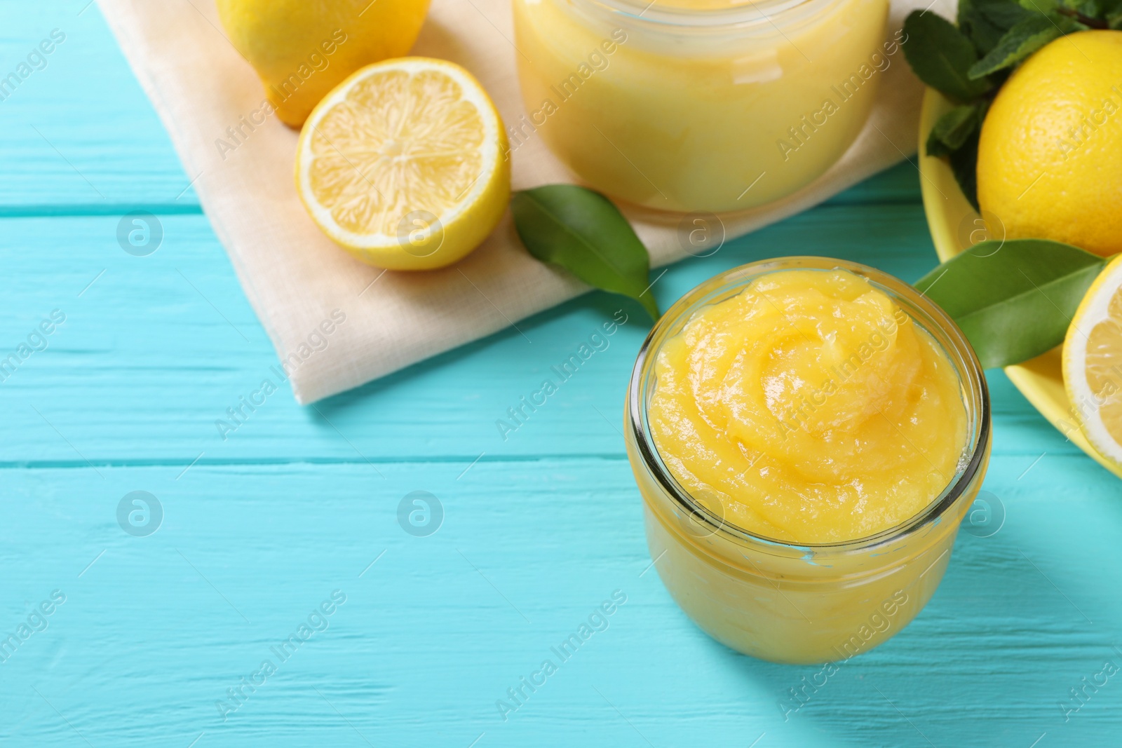 Photo of Delicious lemon curd in glass jar, fresh citrus fruits and green leaves on light blue wooden table, space for text