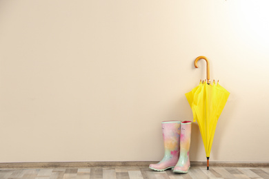 Beautiful yellow umbrella and rubber boots near beige wall. Space for text