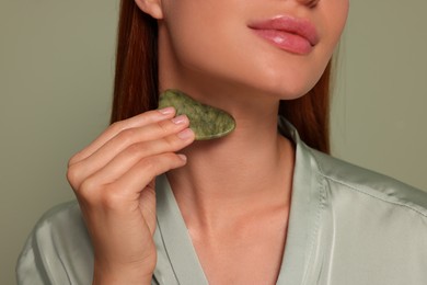 Photo of Young woman massaging her face with jade gua sha tool on green background, closeup