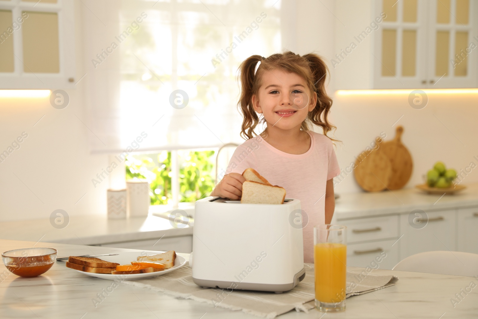 Photo of Cute little girl putting slice of bread into toaster at table in kitchen