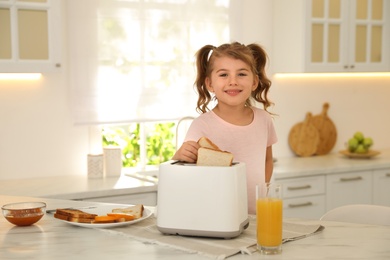 Photo of Cute little girl putting slice of bread into toaster at table in kitchen