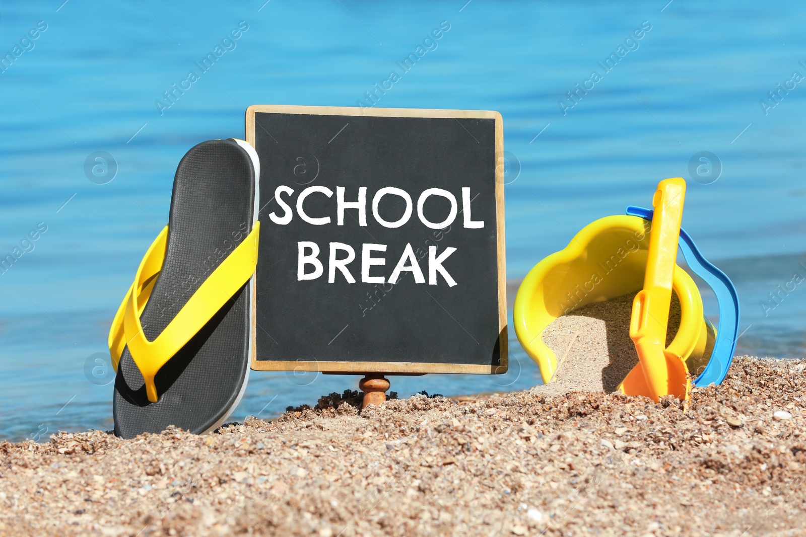 Image of Small black chalkboard with text School Break, beach toys and flip flops on sand near sea. Seasonal holidays 