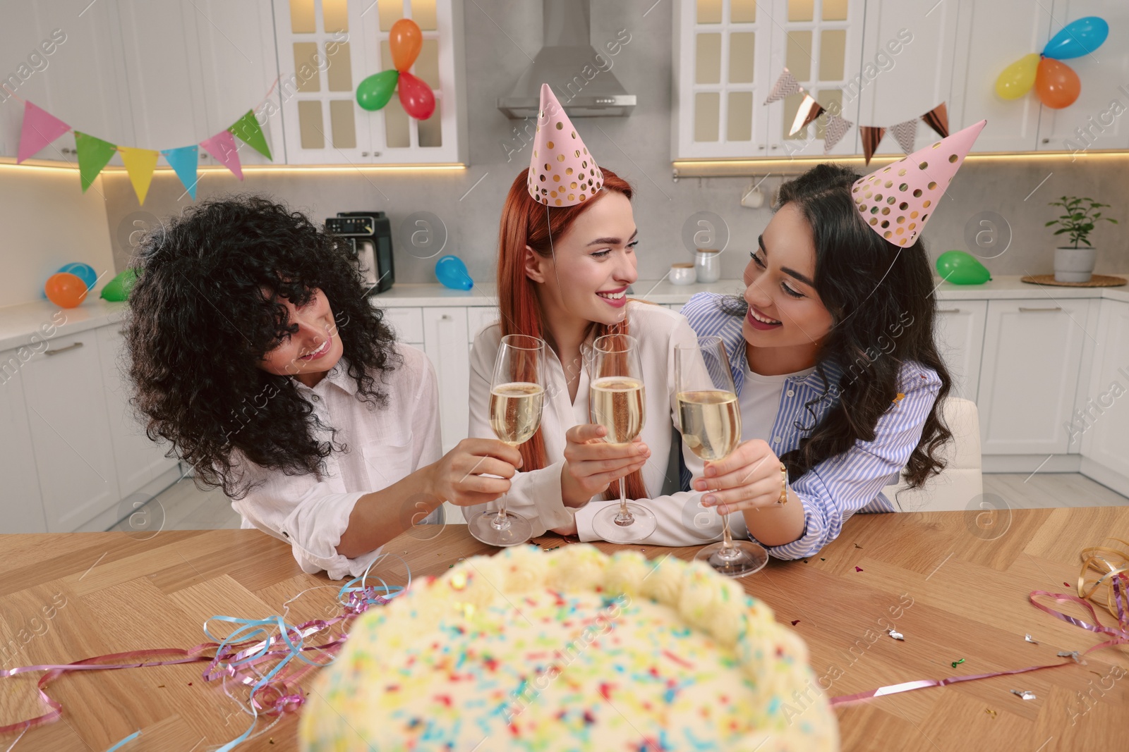 Photo of Happy young women with tasty cake and glasses of sparkling wine celebrating birthday in kitchen