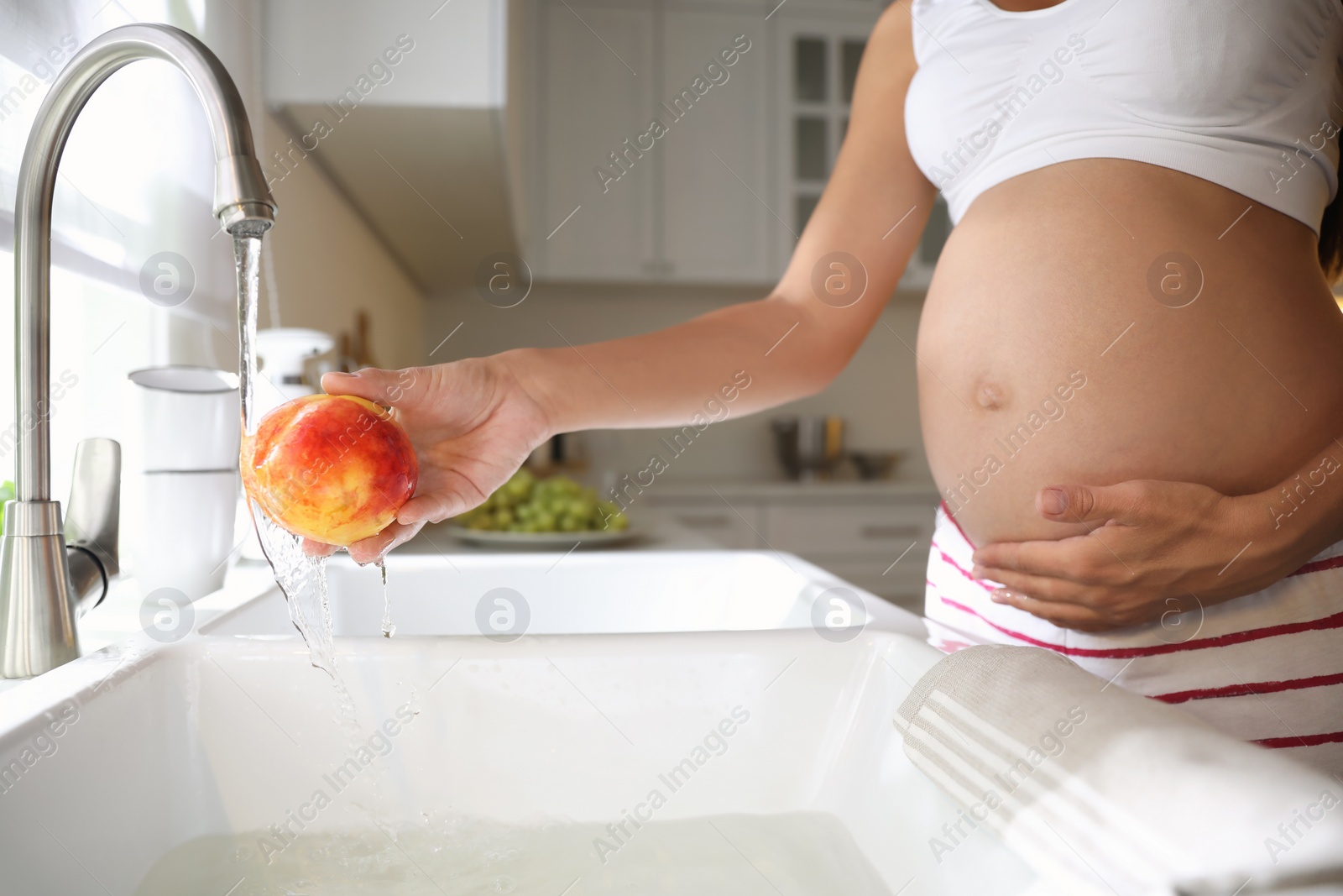 Photo of Young pregnant woman washing fresh sweet peach in kitchen, closeup. Taking care of baby health