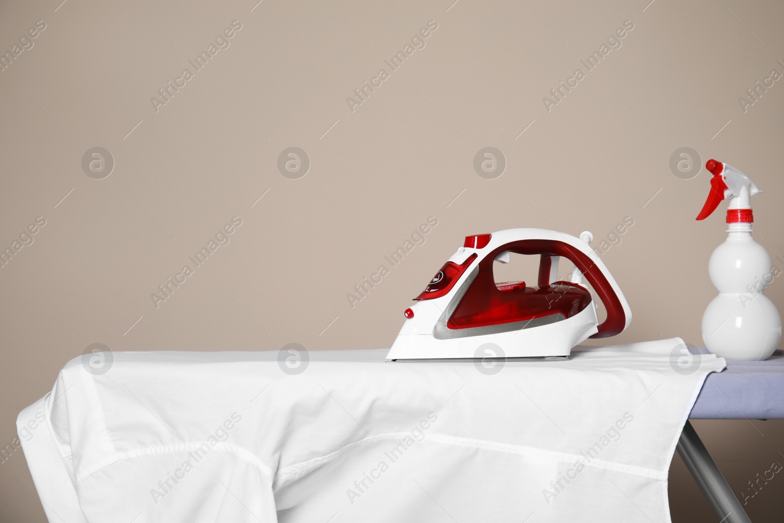 Photo of Modern iron, spray bottle and clean shirt on board against beige background, space for text