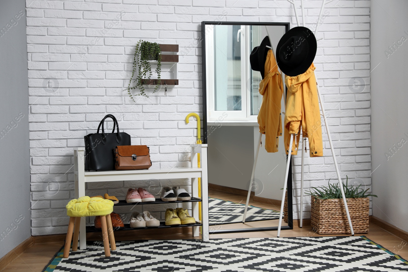 Photo of Stylish hallway interior with coat rack, shoe storage bench and mirror near white brick wall