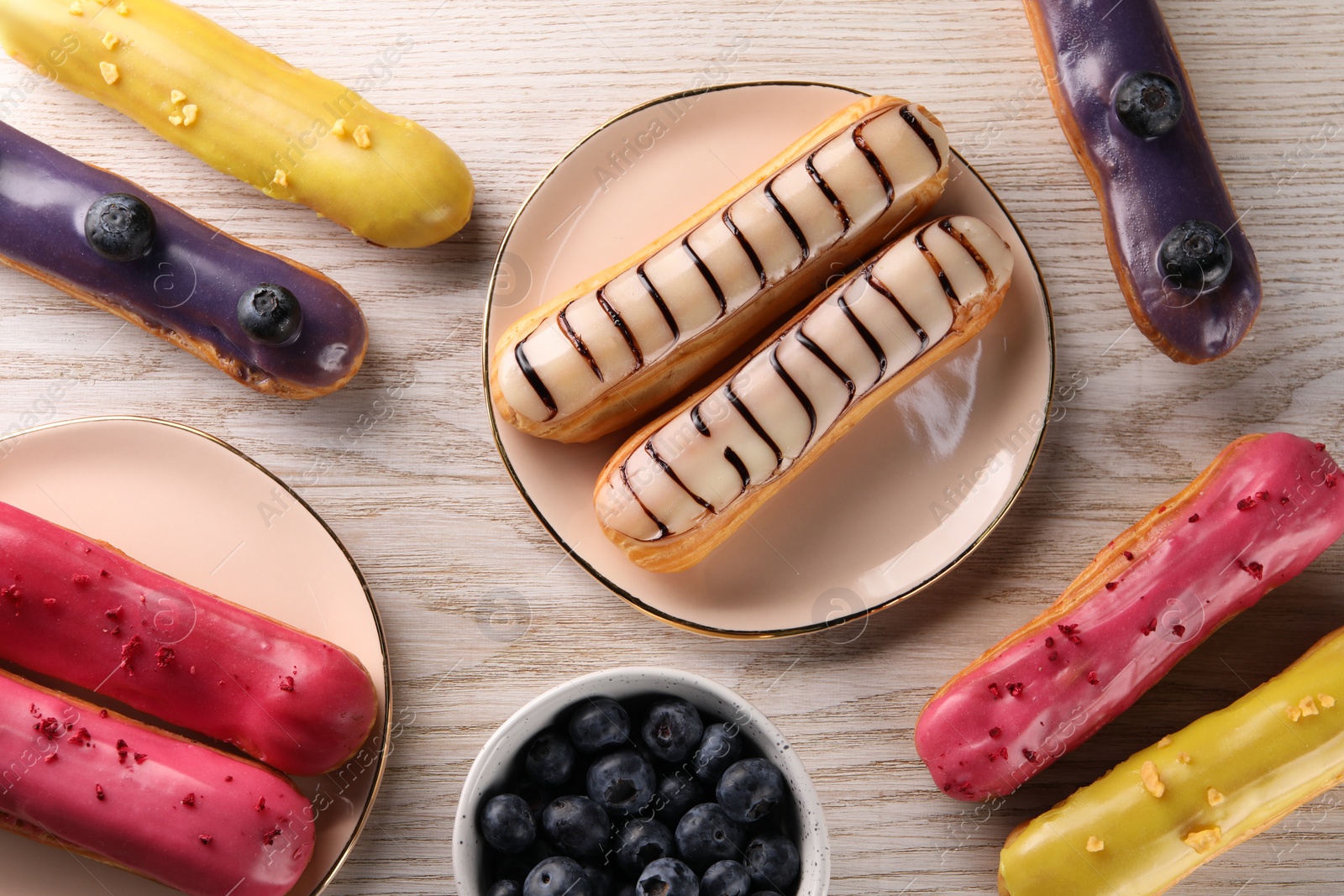 Photo of Different tasty glazed eclairs and blueberries on light wooden table, flat lay