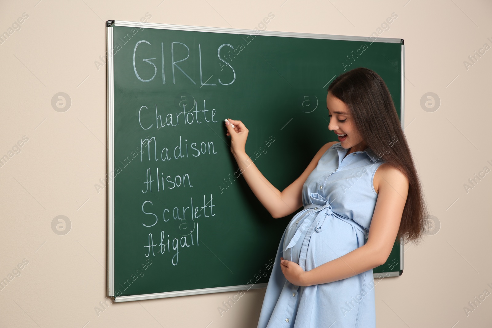 Photo of Pregnant woman writing different baby names on green chalkboard