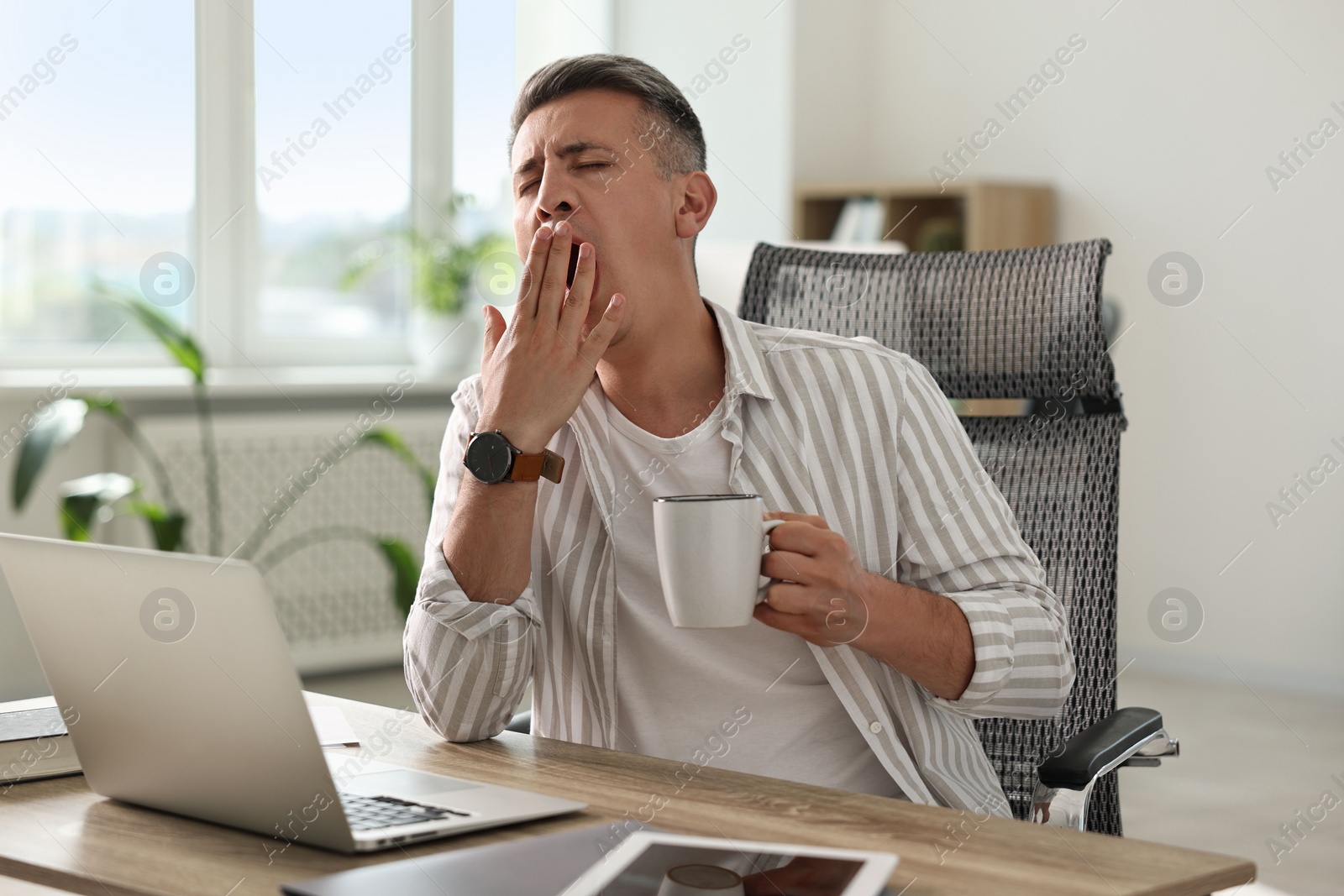 Photo of Man with cup of drink yawning at wooden table in office