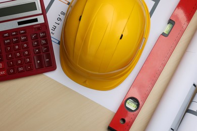 Photo of Construction drawings, safety hat, calculator and bubble level on wooden table, flat lay