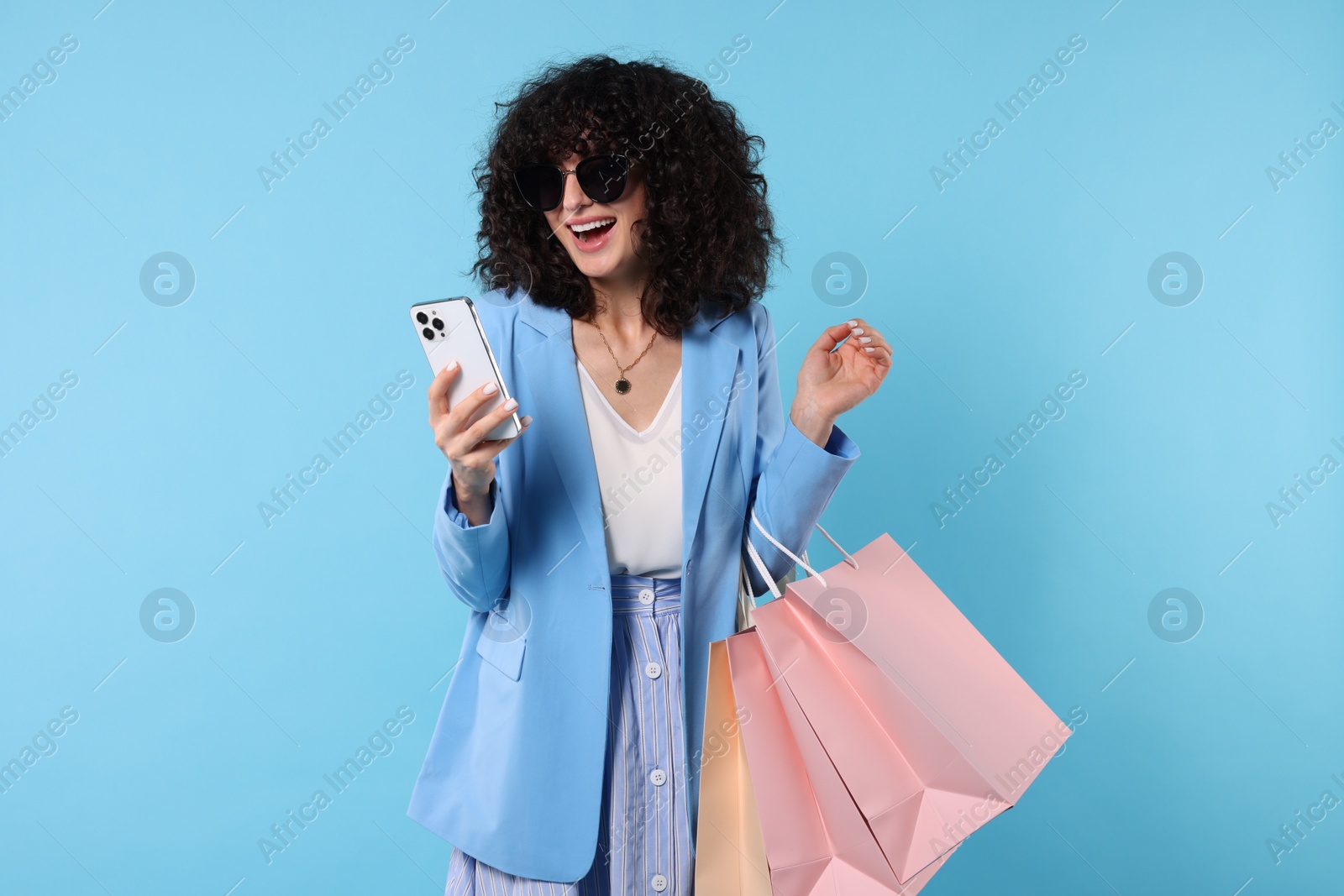 Photo of Happy young woman with shopping bags and modern smartphone on light blue background