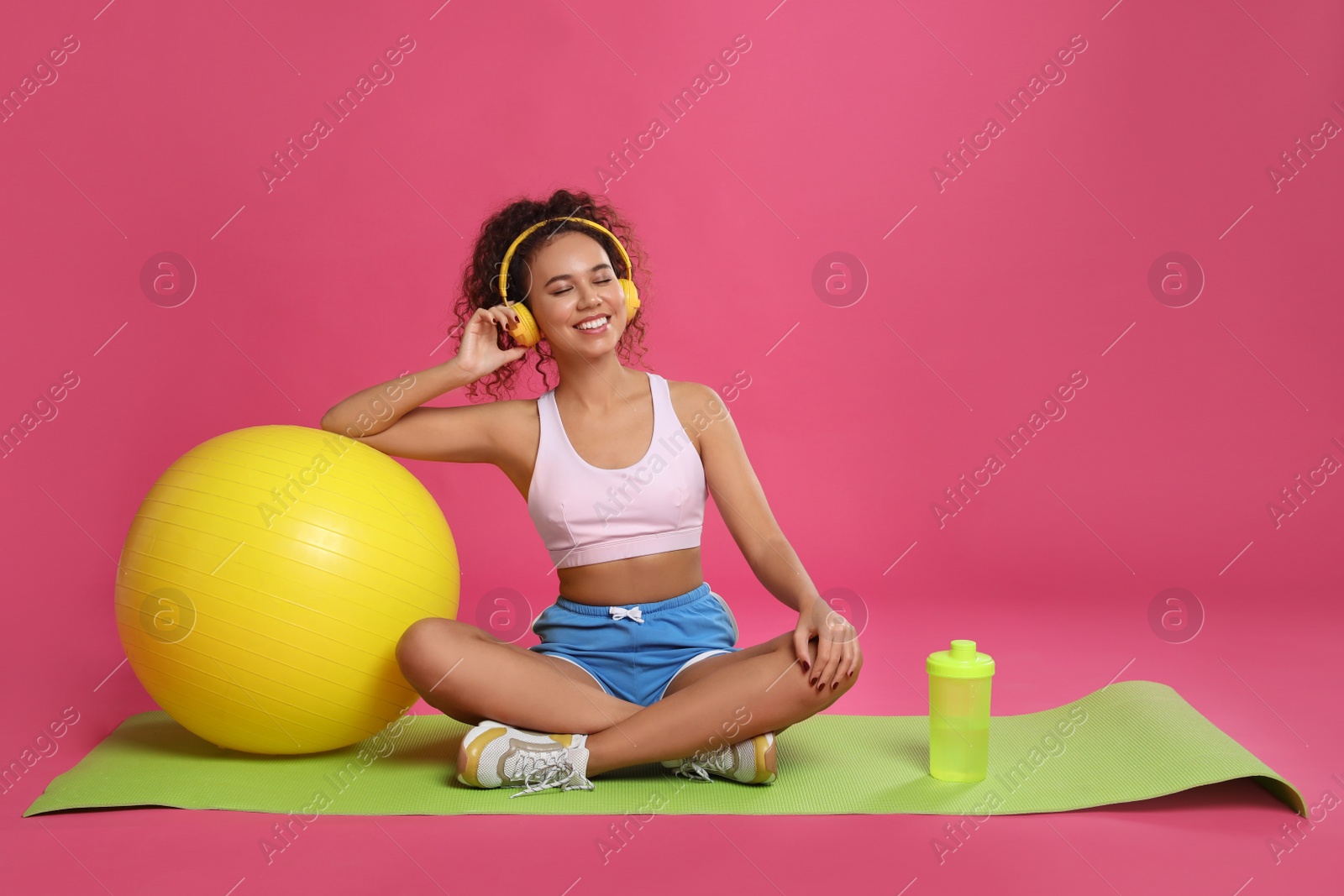 Photo of Beautiful African American woman with headphones  on yoga mat against pink background