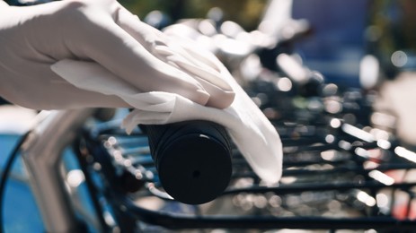 Photo of Woman in latex gloves cleaning bicycle handlebar with wet wipe outdoors, closeup. Protective measures