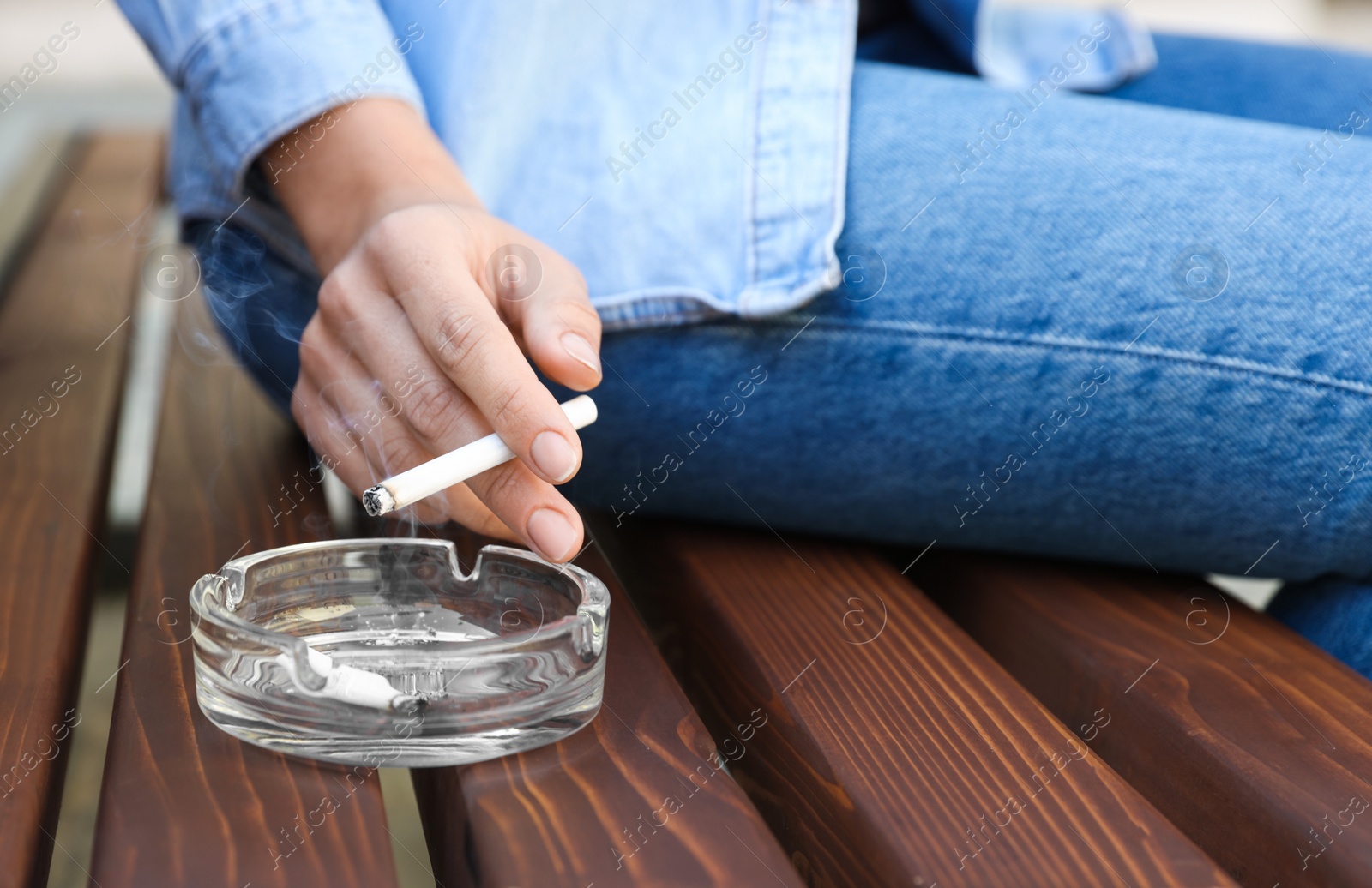 Photo of Woman holding cigarette over glass ashtray on bench outdoors, closeup