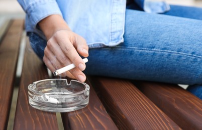 Woman holding cigarette over glass ashtray on bench outdoors, closeup