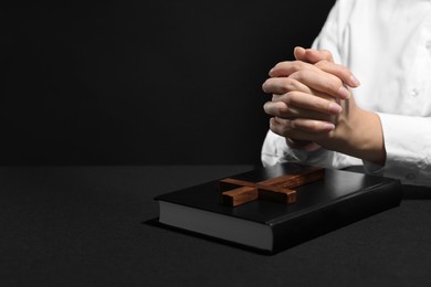 Photo of Woman holding hands clasped while praying at table with Bible against black background, closeup. Space for text