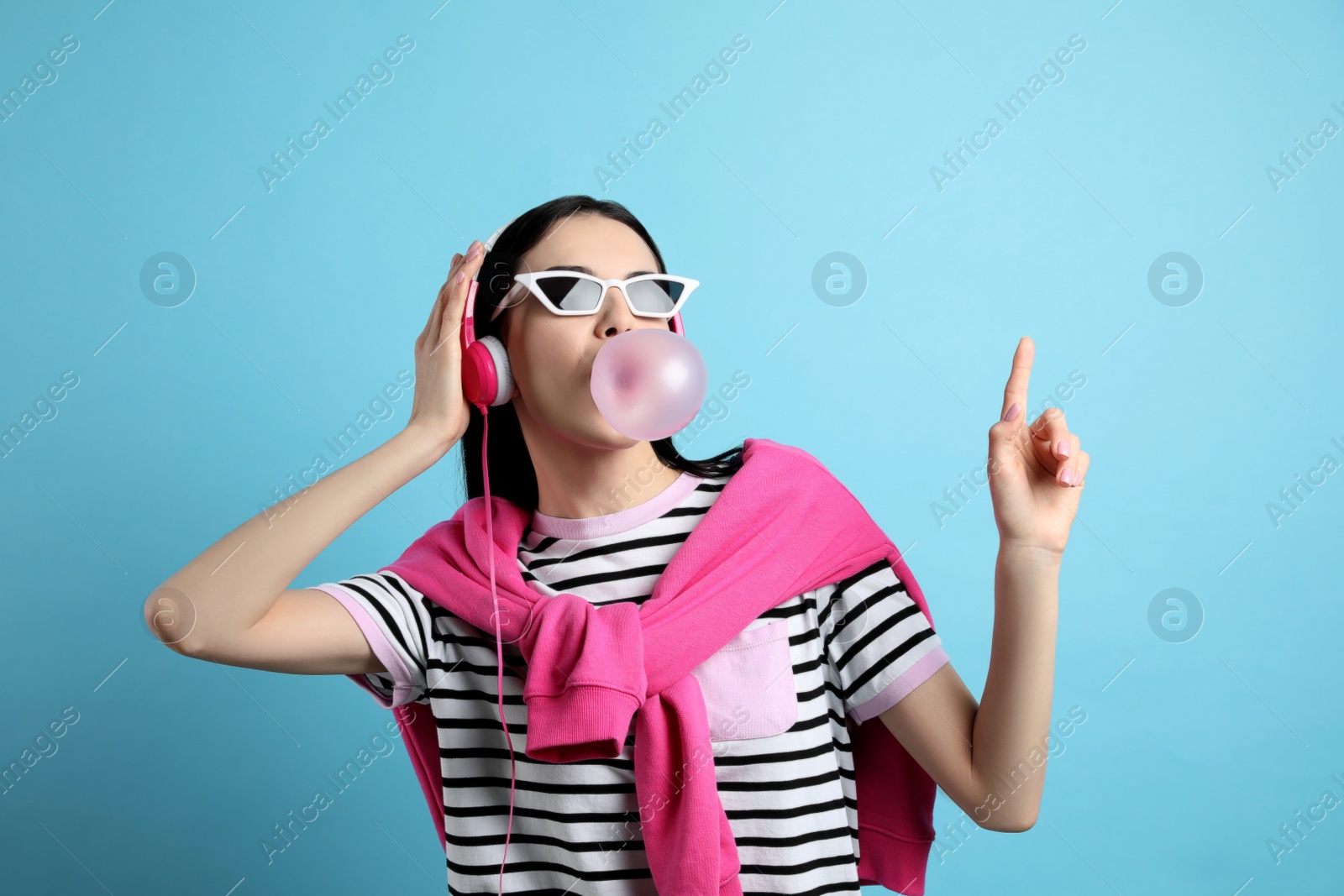 Photo of Fashionable young woman with headphones blowing bubblegum on light blue background