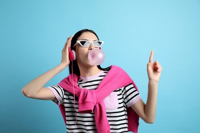 Photo of Fashionable young woman with headphones blowing bubblegum on light blue background