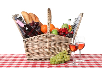 Wicker picnic basket with different products on checkered tablecloth against white background