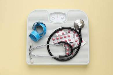 Photo of Scales with weight loss pills, measuring tape, and stethoscope on beige background, top view