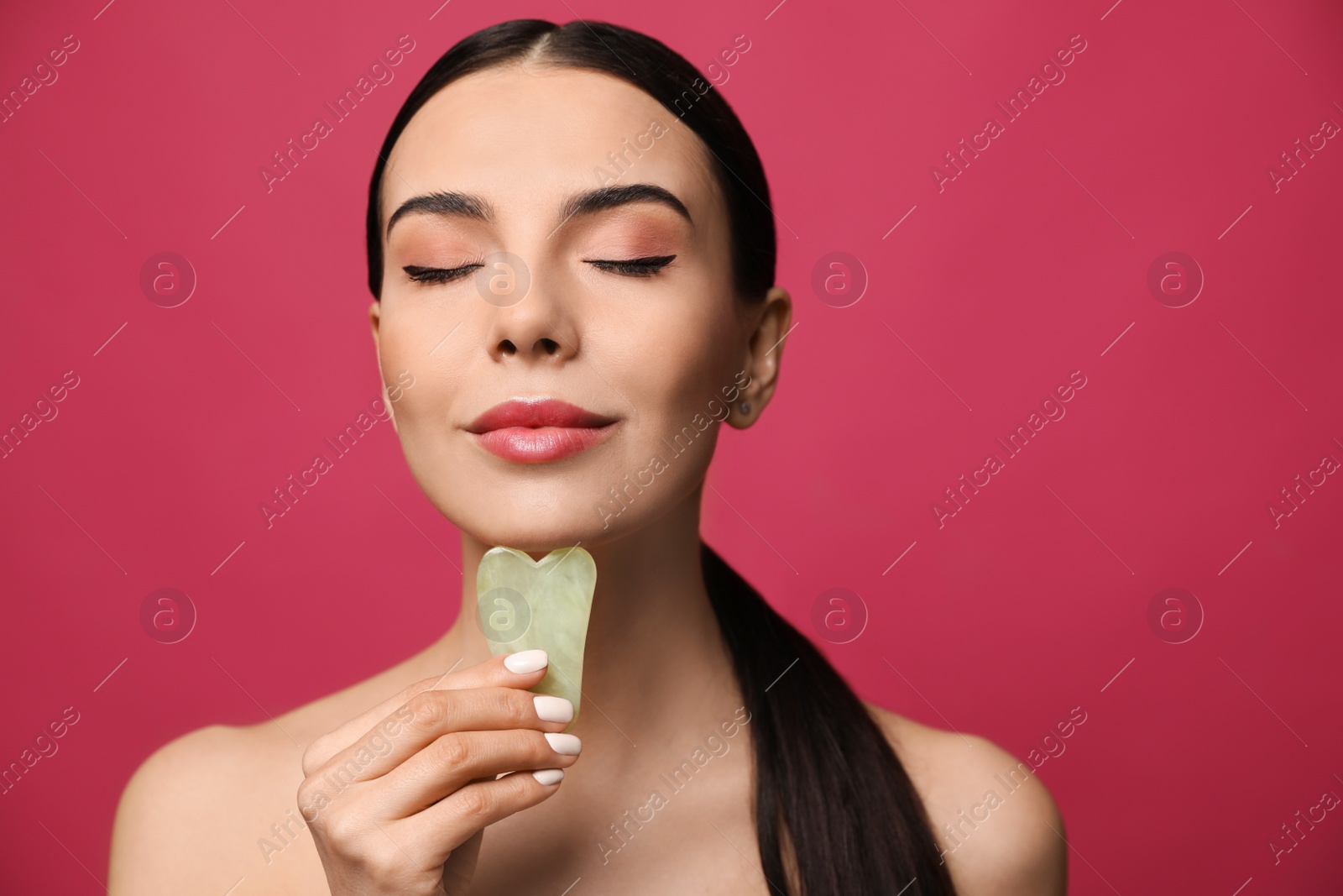 Photo of Beautiful young woman doing facial massage with gua sha tool on pink background