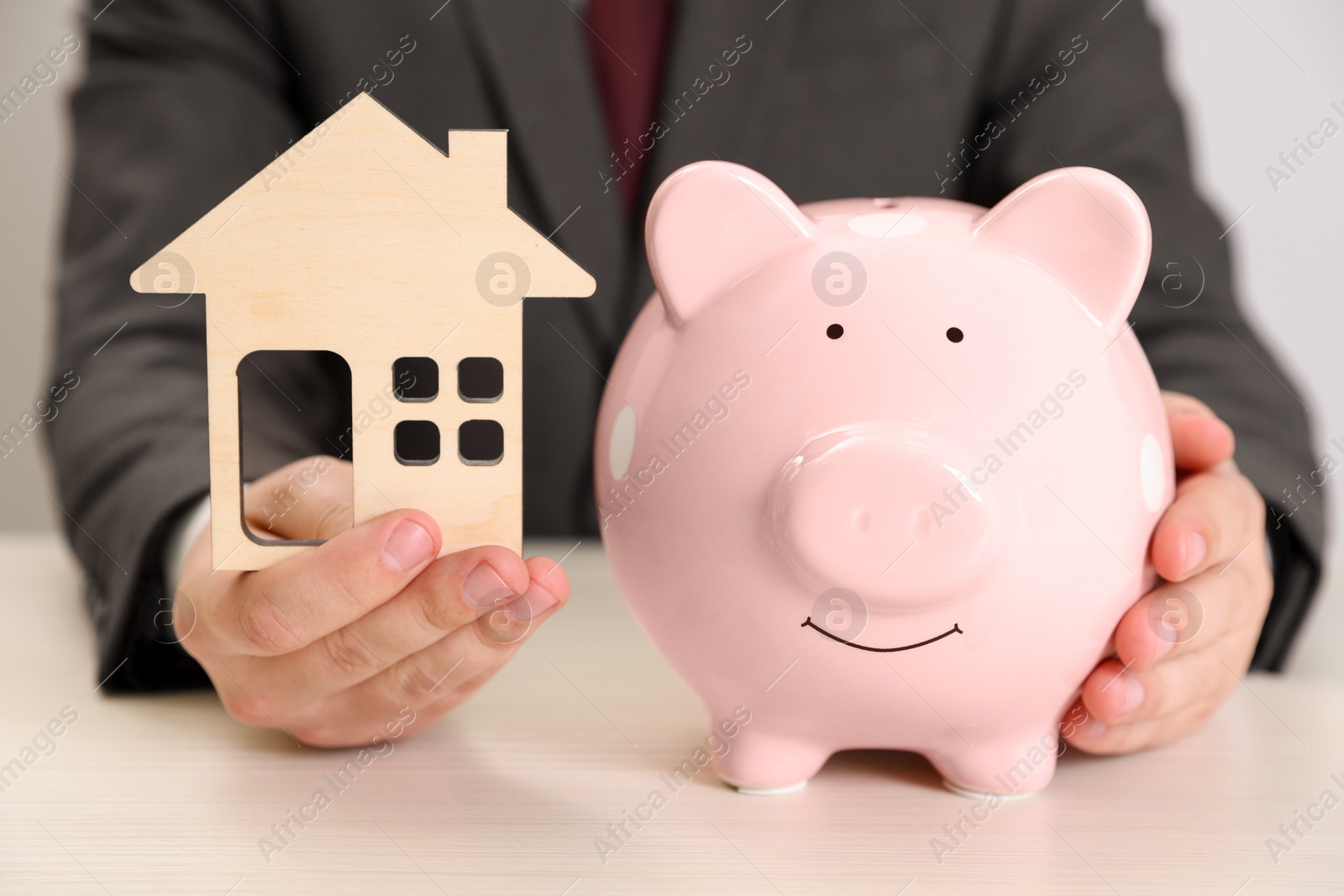 Photo of Man with house model and piggy bank at wooden table, closeup. Saving money concept