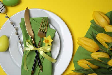 Festive table setting with painted eggs and tulips on yellow background, flat lay. Easter celebration